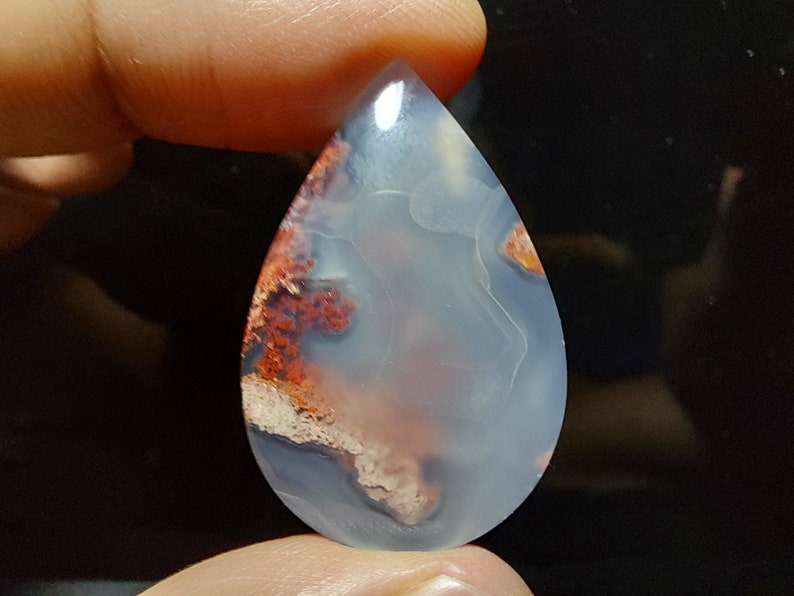 Peut inclure: Un cabochon d'agate en forme de larme avec un motif de couleur blanc, gris et rouge. L'agate a une apparence translucide avec une surface lisse et polie.