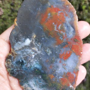 May include: A polished, irregularly shaped stone. It displays a blend of colours, including deep blue, rust-orange, and grey, with touches of green and white. The natural patterns create a unique, abstract design.
