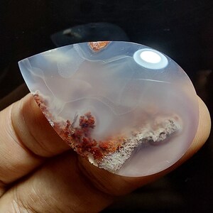 Peut inclure: Un cabochon d'agate en forme de larme avec une base blanche et translucide et des inclusions dendritiques rouges et brunes.