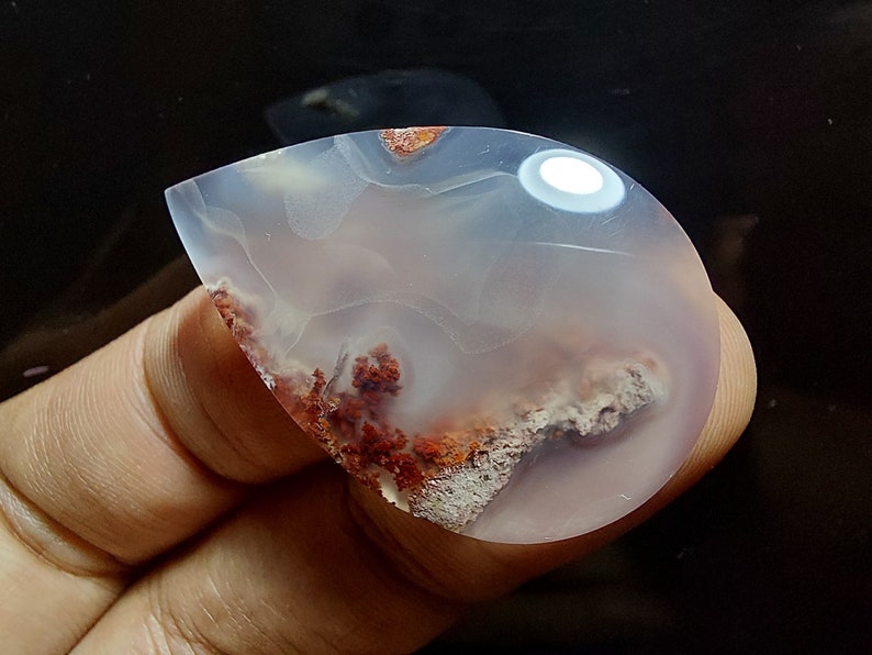 Peut inclure: Un cabochon d'agate en forme de larme avec une base blanche et translucide et des inclusions dendritiques rouges et brunes.