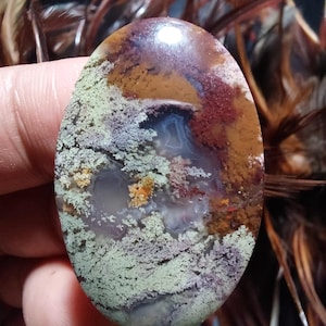 Puede incluir: Una piedra de ágata pulida de forma ovalada con una variedad de colores, incluyendo marrón, rojo, verde y blanco. La piedra tiene un patrón único de remolinos y líneas.