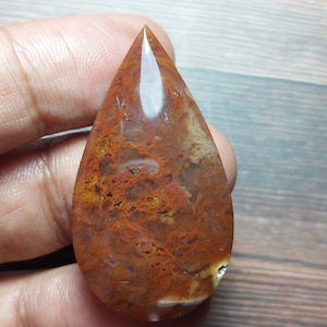 Op de afbeelding: Een traanvormige cabochon van agaat met een rood, oranje en wit patroon. De steen heeft een doorschijnende uitstraling en is gepolijst tot een gladde afwerking.