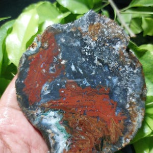 Puede incluir: Una piedra pulida de forma irregular con un llamativo patrón de colores. La piedra presenta una base azul oscuro con vetas rojas, marrones y blancas vibrantes. Los bordes son ásperos y la apariencia general es natural y orgánica.