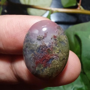 以下が含まれることがあります： 磨かれた光沢のある表面を持つ楕円形の宝石。石は、上部の半透明のピンクがかった紫、緑、赤、白に変化する色の混合を示し、ユニークなパターンを作り出しています。 宝石は指で持たれています。