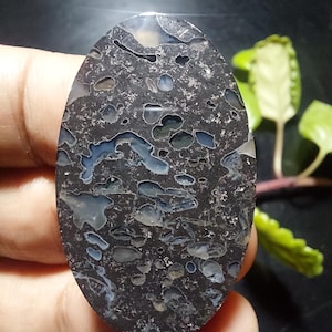 Peut inclure: Une pierre précieuse ovale avec une base gris foncé et des inclusions grises claires et bleues. La surface polie révèle un motif texturé unique. Ce design naturel est idéal pour les bijoux ou l'exposition. Mesure environ 5 cm de long.
