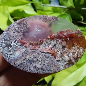 May include: Oval-shaped gemstone with a unique pattern of red, brown, and gray inclusions. The stone has a translucent quality, revealing a landscape-like design. The polished surface highlights the natural variations and textures within the stone.