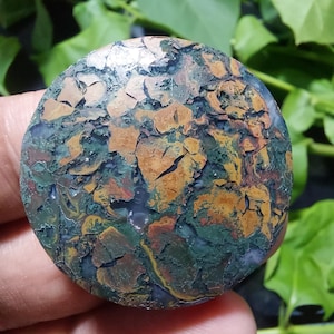 May include: A round, polished stone with a unique, textured surface. The stone features a blend of colors, including green, brown, and hints of red and yellow. The pattern resembles a cracked, earthy landscape. The stone is held in a hand.
