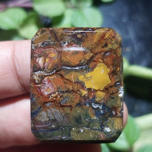 May include: A rectangular gemstone with a polished surface. The stone displays a mix of colours, including browns, oranges, yellows, and greys. The pattern resembles a landscape with various textures and colour variations. The gemstone is held in a hand.