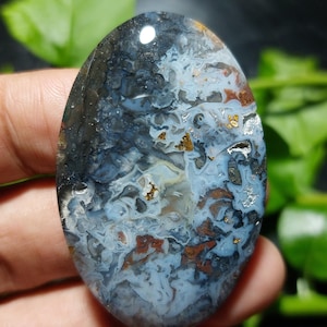 May include: An oval-shaped polished gemstone featuring a complex, swirling pattern of translucent light blue, deep navy, earthy brown, and metallic gold inclusions. The surface is smooth and reflective, showcasing a marbled, multi-layered appearance.