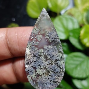 May include: A teardrop-shaped gemstone with a translucent base, featuring moss-like inclusions in green, grey, and purple. The polished surface enhances the natural textures and colours of the stone, creating a unique and organic aesthetic. The stone is held in a hand.