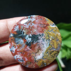 May include: A round, polished gemstone with a vibrant mix of colors. The stone features a blend of red, yellow, black, and white hues, creating a unique and natural pattern. The gemstone is held in a hand, showcasing its size and detail.
