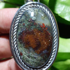 May include: A silver pendant featuring a green, brown and red agate stone set within a rope-like border.