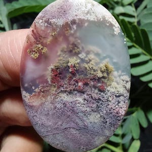 May include: A polished oval agate cabochon with a unique landscape pattern. The stone features shades of white, gray, pink, red, and green. The agate has a natural, organic look.