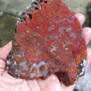 Puede incluir: Una piedra natural en bruto con un llamativo patrón rojo y marrón, acentuado por vetas blancas y azules. La piedra tiene una superficie texturizada y una forma irregular, mostrando su belleza cruda y orgánica. Una pieza decorativa única.