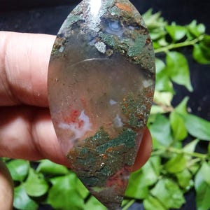 May include: A polished, elongated gemstone with a pointed tip. The stone features a mix of colors, including green, brown, red, and white, with a translucent quality. The gemstone is held in a hand, with green foliage in the background.