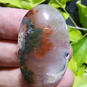 Op de afbeelding: Een ovale edelsteen met een gepolijst oppervlak. De steen heeft een mix van kleuren, waaronder doorschijnend wit, roze, groen en oranje tinten. Het patroon lijkt op een landschap met organische vormen en texturen. De edelsteen wordt in een hand gehouden.