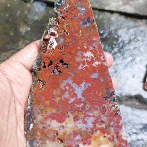 May include: A triangular-shaped, natural stone slab with a rough, uneven edge. The stone features a vibrant mix of red, orange, white, and blue hues, creating a unique, marbled appearance. The stone is held in a hand.