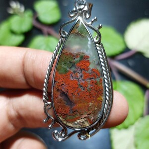 Può includere: Ciondolo a goccia con cornice color argento. Il ciondolo presenta una pietra lucida con motivi rossi, verdi e marroni. La cornice ha un design a corda intrecciata e riccioli decorativi nella parte superiore.