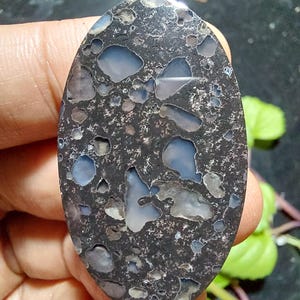 Peut inclure: Une pierre précieuse de forme ovale avec une base gris foncé et des motifs irréguliers de bleu clair et de blanc. La surface polie révèle un aspect moucheté unique. La pierre est tenue dans une main, mettant en valeur sa taille et sa forme.