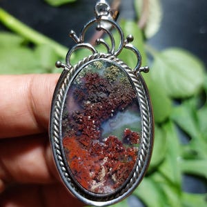 Puede incluir: Colgante ovalado con marco plateado. El colgante presenta una piedra natural con un patrón similar a un paisaje en tonos rojos, verdes y marrones. El marco tiene un diseño de cuerda trenzada alrededor de la piedra y adornos decorativos en la parte superior.