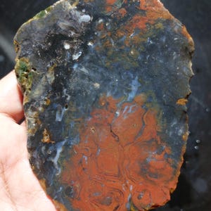 Peut inclure: Une dalle de pierre polie de forme irrégulière avec un motif saisissant. La pierre présente un mélange de couleurs, notamment du noir profond, du rouge rouille et des touches de bleu et de vert. La surface présente des motifs tourbillonnants complexes.