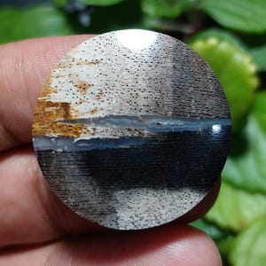 May include: A round, polished gemstone with a unique pattern. The stone features a mix of colors, including black, white, brown, and a hint of blue. The surface has a textured appearance, with visible lines and small dots. The gemstone is likely used for jewelry or decorative purposes.