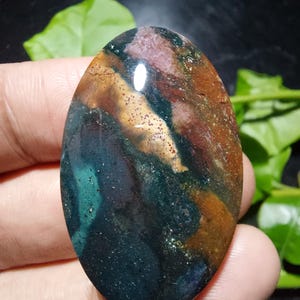 Könnte beinhalten: Ein oval geschliffener Edelstein mit einem komplexen Muster aus tiefem Waldgrün, erdigem Braun und gedämpften rötlich-rosa Tönen. Die Oberfläche weist ein glattes, reflektierendes Finish mit subtilen Variationen in der Farbdichte auf.