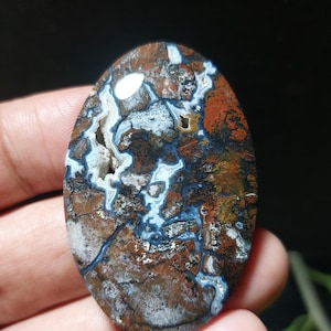 Peut inclure: Pierre précieuse polie de forme ovale avec un motif unique. La pierre présente un mélange de couleurs, notamment le marron, le blanc, le bleu et l'orange. Le motif complexe ressemble à un paysage naturel ou à de l'art abstrait. La pierre est tenue dans une main.