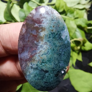 Op de afbeelding: Een ovale edelsteen met een uniek kleurenpatroon. De steen heeft een mix van diepblauwgroen, bordeauxrood en bruin, wat een gemarmerd effect creëert. Het gepolijste oppervlak reflecteert licht en benadrukt de ingewikkelde details van de natuursteen. Ongeveer 5 cm lang.