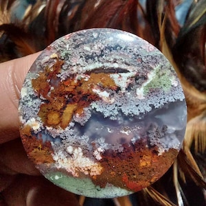 Puede incluir: Una piedra de ágata redonda y pulida con un patrón de color marrón, blanco, gris y verde en espiral. La piedra tiene un aspecto natural y orgánico.