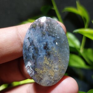 May include: An oval-shaped gemstone with a unique pattern of blue, gray, and gold hues. The stone has a polished surface, and the intricate veining creates a natural, artistic design. The gemstone is held in a person's hand, with green foliage in the background.