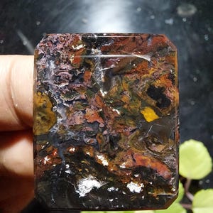 Op de afbeelding: Een rechthoekige edelsteen met een gepolijst oppervlak. De steen heeft een mix van kleuren, waaronder bruin, zwart, oranje, geel en wit, waardoor een gemarmerd effect ontstaat. De edelsteen wordt in een hand gehouden.