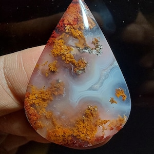 May include: A teardrop-shaped agate cabochon with a white, orange, and red color pattern. The agate has a translucent appearance with a swirling pattern of white and orange.