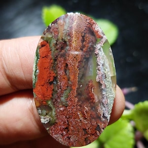 Puede incluir: Una gema ovalada con un aspecto natural y terroso. La piedra presenta una mezcla de tonos rojos, verdes y marrones, con una superficie texturizada y moteada. Los colores se distribuyen en un patrón que recuerda a un paisaje.