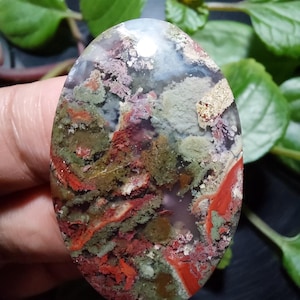 Pode incluir: Um close-up de um cabochão de ágata verde, vermelho e branco com um padrão de redemoinho. O cabochão é oval e tem uma superfície lisa e polida.