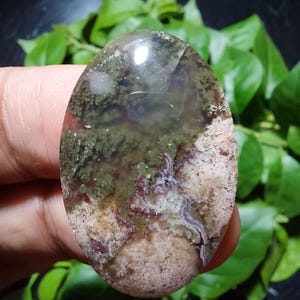 Puede incluir: Una gema ovalada con una superficie translúcida. La piedra presenta una mezcla de colores, incluyendo verde, marrón y beige, con algunas áreas casi transparentes. La gema se sostiene sobre un fondo de hojas verdes.