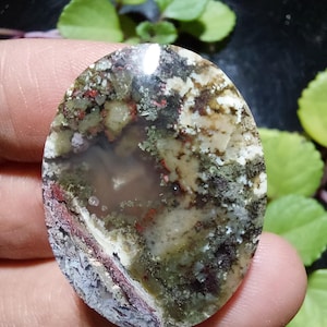 May include: An oval-shaped gemstone with a unique pattern of colors. The stone features a mix of green, brown, red, and white hues, creating a natural, earthy appearance. The polished surface reflects light, highlighting the intricate details within the stone.