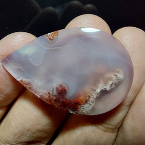 Peut inclure: Un cabochon d'agate en forme de larme avec une base blanche et rose clair et des inclusions brunes et rouges. La pierre est translucide et a une surface lisse et polie.