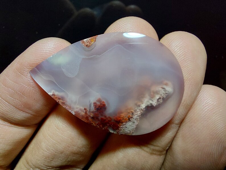 Peut inclure: Un cabochon d'agate en forme de larme avec une base blanche et rose clair et des inclusions brunes et rouges. La pierre est translucide et a une surface lisse et polie.