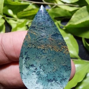 May include: A teardrop-shaped moss agate gemstone featuring a polished surface with intricate inclusions of deep forest green, sage, and earthy brown moss-like patterns throughout the stone, displaying a smooth, rounded edge and a pointed top.