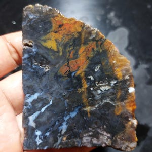 May include: A polished, irregularly shaped stone with a striking pattern of colours. The stone features a mix of dark grey, black, orange, yellow, and white hues, with intricate veining and marbling. The edges are rough, showcasing the natural texture of the stone.