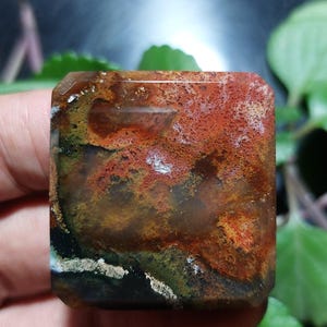 Peut inclure: Une pierre précieuse de forme carrée aux couleurs riches et terreuses. La pierre présente un mélange de teintes rouges, oranges, brunes, vertes et blanches, créant un effet marbré. La surface polie reflète la lumière, soulignant les motifs naturels.