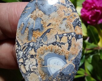 Puede incluir: Un cabochón ovalado pulido de ágata colorida con un patrón de remolino de marrón, gris y blanco. La piedra tiene una textura moteada y un acabado brillante.