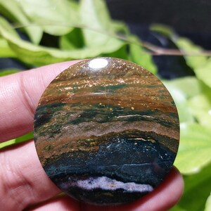 May include: A circular polished gemstone cabochon featuring horizontal bands of earthy colors including deep forest green, dark brown, tan, and a thin layer of white and purple near the bottom edge, with a smooth, reflective, and glossy surface finish.