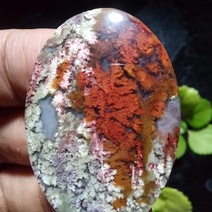 Puede incluir: Una piedra preciosa ovalada con un patrón único de inclusiones rojas, blancas y marrones. La superficie pulida de la piedra presenta una mezcla de colores y texturas. El diseño evoca un paisaje natural.
