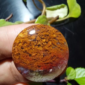 May include: A round, polished gemstone with a layered appearance. The top layer is a rich, rusty orange with intricate patterns. Below, a band of red and white leads to a translucent base with hints of green. The stone is held in a hand.