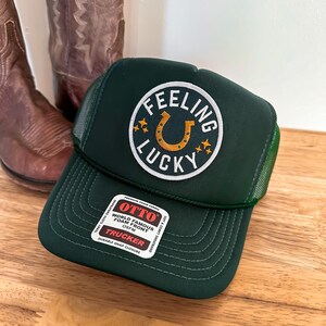 Può includere: Cappellino da camionista verde con una toppa bianca che dice "Feeling Lucky" con un ferro di cavallo dorato al centro. Il cappello ha una parte posteriore in rete nera e una chiusura a scatto nera.