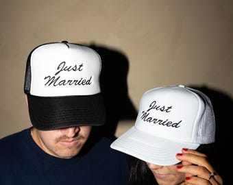 Op de afbeelding: Twee witte trucker caps met de tekst "Just Married" in zwart. Eén cap heeft een zwarte klep en mesh achterkant, de andere een witte klep en mesh achterkant. Ideaal voor een pasgetrouwd stel.