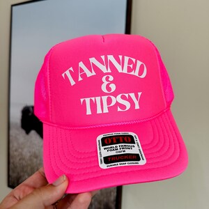 May include: A bright pink trucker hat with the words "TANNED & TIPSY" in white block letters. The hat has a mesh back and a curved brim. The hat also has a tag that says "WORLD FAMOUS FOAM FRONT".