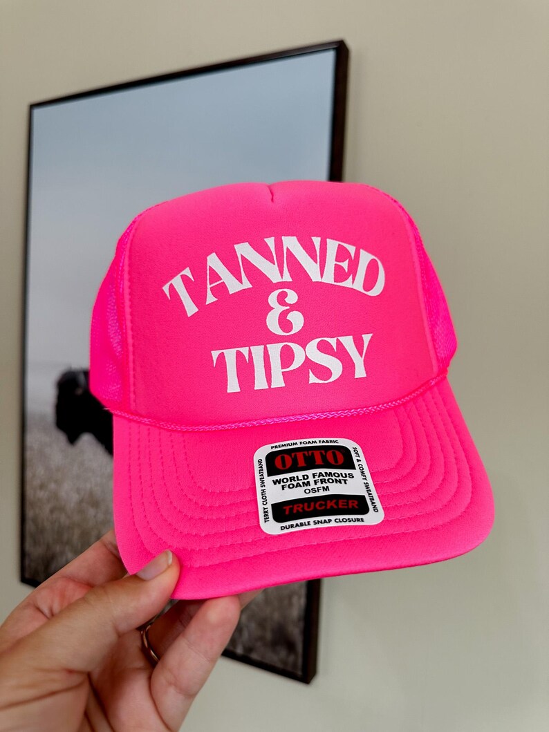 May include: A bright pink trucker hat with the words "TANNED & TIPSY" in white block letters. The hat has a mesh back and a curved brim. The hat also has a tag that says "WORLD FAMOUS FOAM FRONT".